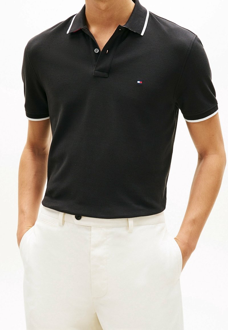 Homme portant un polo noir à manches courtes avec des bordures blanches et un pantalon blanc, les mains dans les poches.