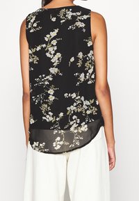 Blouse noire sans manches avec un imprimé floral composé de fleurs blanches et jaunes, en tissu léger, avec un ourlet arrondi et une texture lisse.