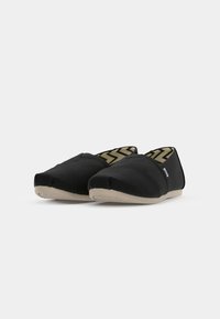Sorte slip-on sko laget av lerret med en teksturert overflate, med en rund tå, beige gummisåle og et innvendig fôr med zigzag-mønster.