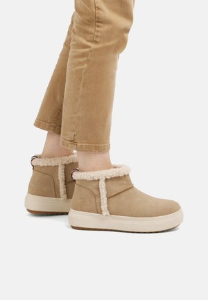 Person iført tan ankelstøvler med hvid pelskant og beige bukser, stående mod en hvid baggrund.