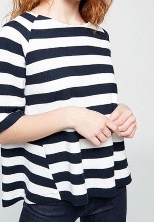 Vrouw draagt een losse zwart-wit horizontaal gestreepte top met ellebooglange mouwen, handen zachtjes samengevouwen voor zich.