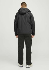 Zwarte jas met capuchon, met een gladde textuur en ritssluiting, gecombineerd met donkere cargo-broeken en sneakers in een neutrale kleurenpalet.