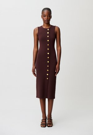 Femme portant une robe midi marron sans manches avec une rangée verticale de boutons dorés sur le devant, associée à des talons noirs à lanières.