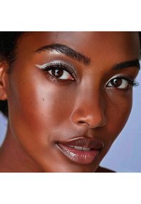 Gros plan sur un visage avec une peau brune lisse et chaude ; des cils foncés et recourbés ; des sourcils bien définis ; et un eyeliner argenté qui accentue les yeux.