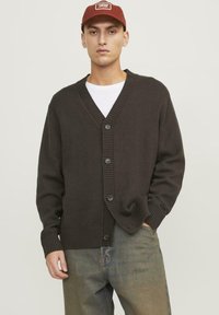 Cardigan lavorato a maglia marrone scuro con scollo a V, chiusura a cinque bottoni, polsini e orlo a costine. Indossato sopra una maglietta bianca e abbinato a jeans blu.