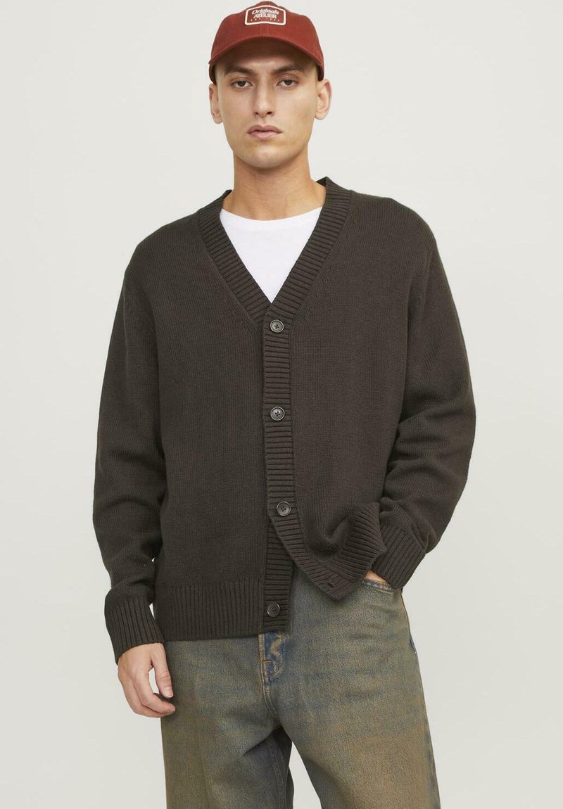 Cardigan lavorato a maglia marrone scuro con scollo a V, chiusura a cinque bottoni, polsini e orlo a costine. Indossato sopra una maglietta bianca e abbinato a jeans blu.