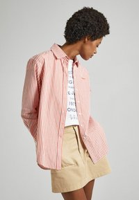 Maglietta a righe rosse e bianche con colletto a camicia, abbinata a una gonna beige con tasche a patta. Tessuto texture, vestibilità comoda.
