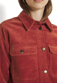 Mujer con chaqueta de pana roja con grandes bolsillos delanteros y botones plateados, mostrando el cuello y los hombros.