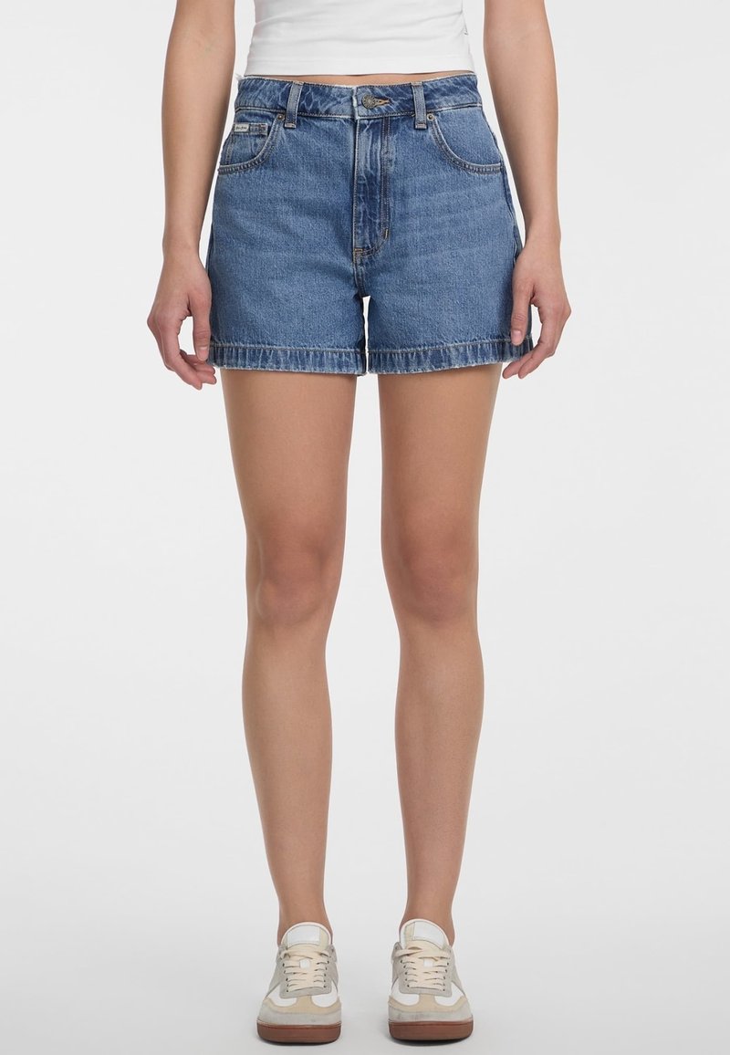 Femme portant un short en denim bleu taille moyenne avec poches frontales, fermeture à boutons et zip, associé à des baskets blanches et un top court blanc.