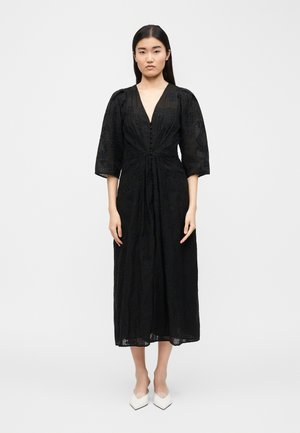 Femme debout portant une robe midi noire avec des manches trois-quarts bouffantes et des escarpins blancs à bout pointu, sur un fond uni.