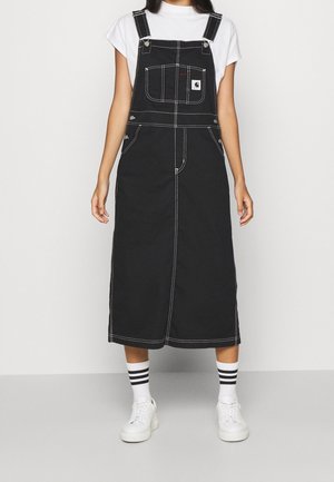 Woman wearing a black denim pinafore dress with white stitching, a white T-shirt, striped socks, and white sneakers, standing on a gray floor.