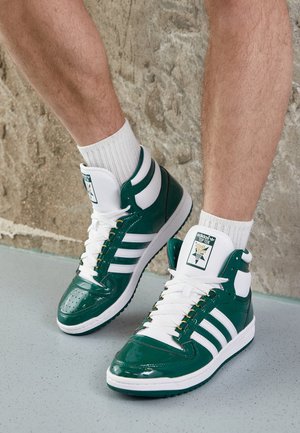Person iført grønne og hvide Adidas Top Ten høje sneakers med hvide sokker, stående mod en struktureret væg.