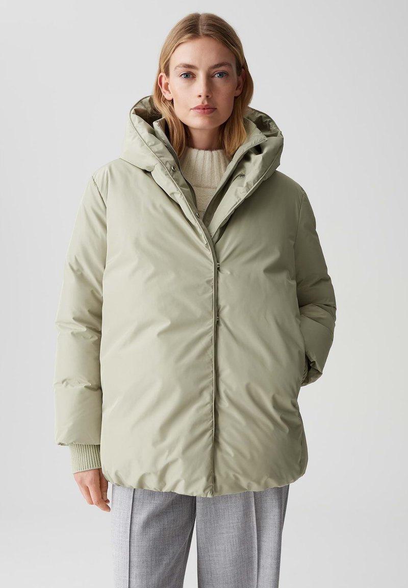 Giacca puffer verde chiaro con un grande cappuccio, zip frontale e dettagli a costine sui polsini. Presenta una texture liscia e lucida e una vestibilità ampia.