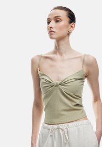 Top sin mangas de color verde claro, texturizado, con tirantes finos y un detalle de nudo en la parte delantera, combinado con pantalones de tiro medio de color claro con cordones.
