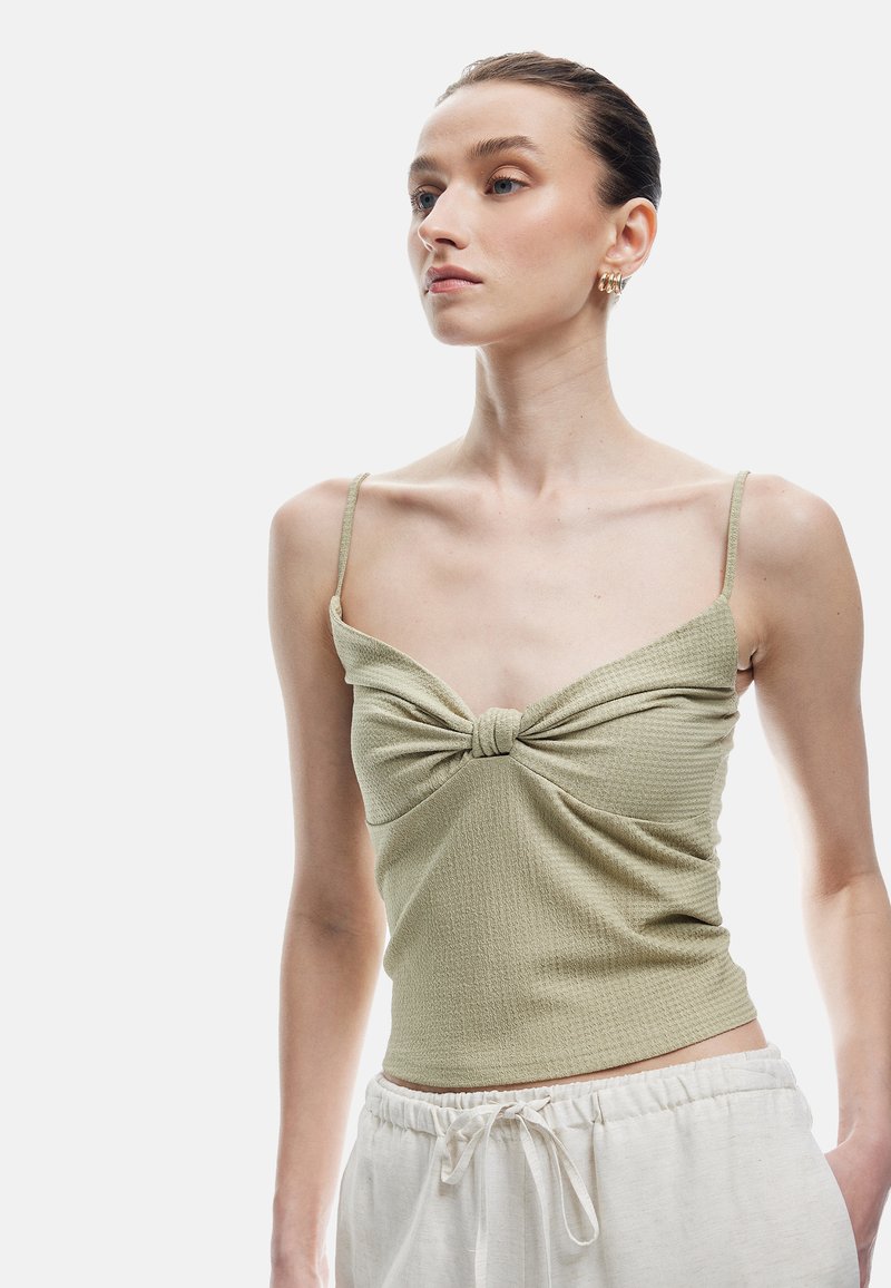 Top sin mangas de color verde claro, texturizado, con tirantes finos y un detalle de nudo en la parte delantera, combinado con pantalones de tiro medio de color claro con cordones.
