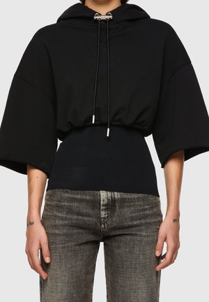 Sweat à capuche court noir avec manches larges et cordons, associé à un jean gris délavé taille haute porté par une personne debout devant un fond neutre.