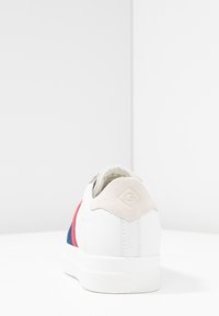 Białe sneakersy z szarym zamszowym piętkiem, czerwonymi i niebieskimi akcentami z tkaniny, teksturowaną podeszwą oraz detalem logo na pięcie. Minimalistyczny design.