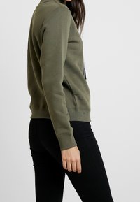 Sudadera verde oliva con cuello redondo, puños y dobladillo acanalados. Cuenta con un bolsillo lateral y un corte holgado. Los materiales parecen suaves y con textura.