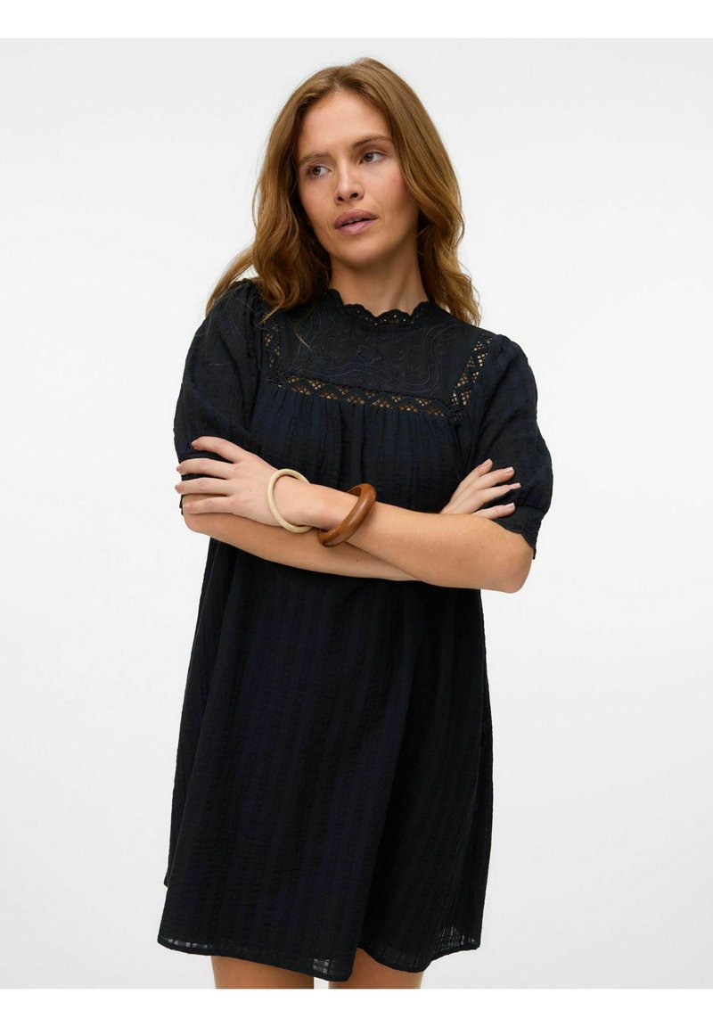 Črna obleka s kratkimi napihnjenimi rokavi, teksturirana tkanina, čipkasti detajli na korpusu in valovit izrez. Dodatki vključujejo lesene zapestnice.