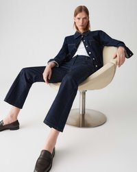 Veste en denim foncé et pantalon avec des coutures visibles, associé à un haut blanc et des mocassins foncés, assis dans une chaise ronde en métal.