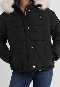 Svart quiltad jacka med pälsbeklädd luva, blixtlås framtill och tryckknappar, bärs med blå jeans.