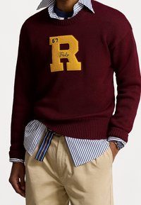Mörkröd stickad tröja med en stor gul "R"-applikation, lagd över en blå och vit randig skjorta. Ljus khaki-byxor och en klocka syns.