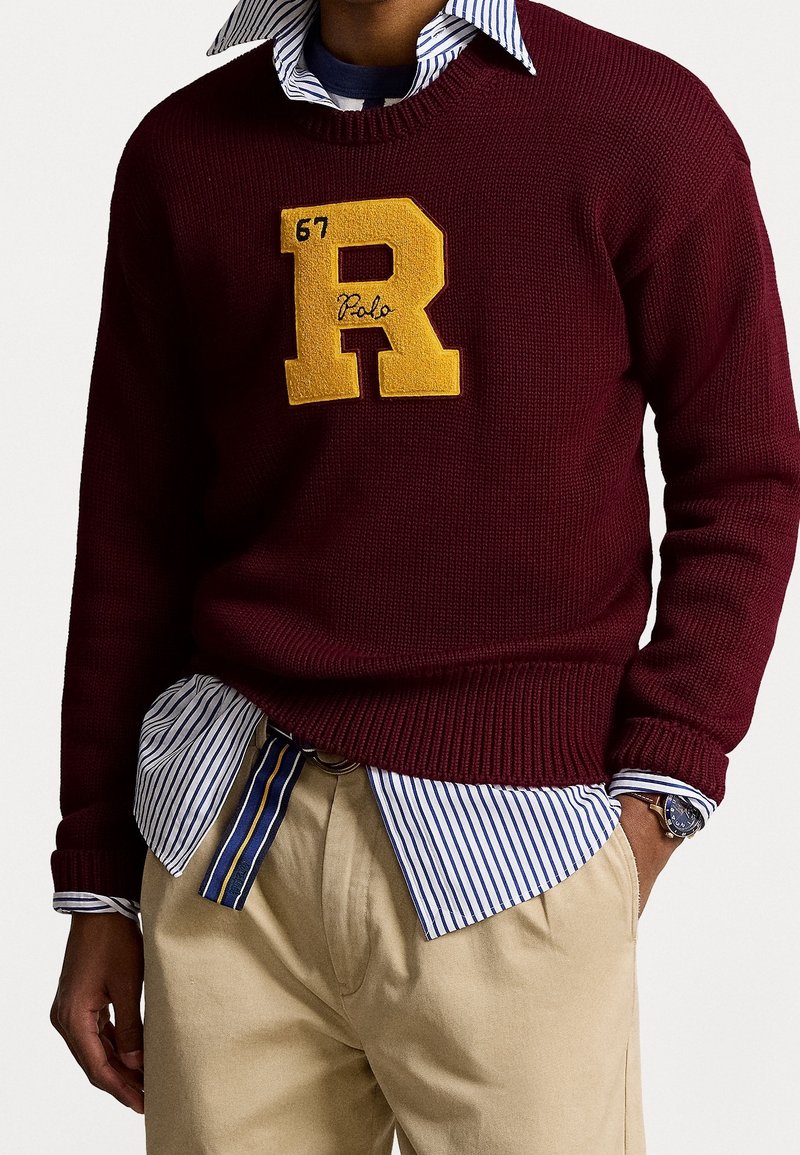 Mörkröd stickad tröja med en stor gul "R"-applikation, lagd över en blå och vit randig skjorta. Ljus khaki-byxor och en klocka syns.