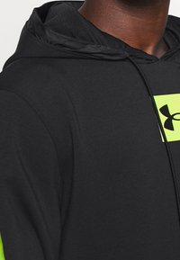 Felpa nera con una texture liscia, caratterizzata da un logo rettangolare giallo-verde e un cordino. Il design include un cappuccio comodo.