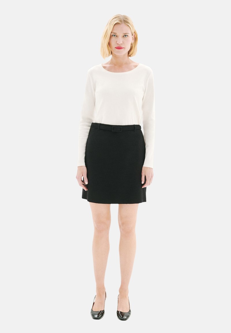 Un haut à manches longues blanc associé à une jupe noire taille haute avec une ceinture. La tenue est complétée par des escarpins noirs.