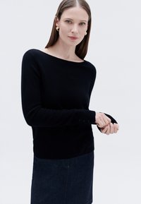 Schwarzer Strickpullover mit weitem Bootsausschnitt und langen Ärmeln. Verfügt über knopfbare Manschetten und eine schlanke Passform, kombiniert mit einem dunklen Jeansrock.