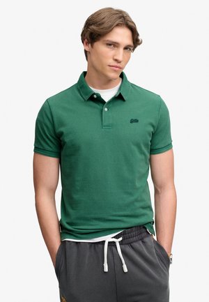 Młody mężczyzna o jasnobrązowych włosach ma na sobie zieloną koszulkę polo z krótkim rękawem na białym T-shircie oraz ciemnoszare spodnie ze sznurkiem.