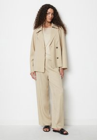 Beige ülikond, millel on kahekordne väljaklapp ja laiad püksid, kokku sobitatud heleda topi ja mustade sandaalidega neutraalsel taustal.