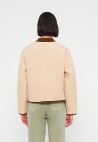 Μπεζ μπουφάν με καπιτονέ, καφέ γιακά και φάσα, με κοντό σχέδιο και μακριά μανίκια, φορεμένο πάνω από πράσινο παντελόνι.