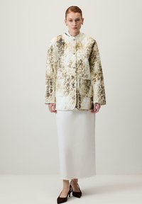 Egy nő bélelt, gombos dzsekit visel, aranybarna és fekete batikolt mintával, fehér bokáig érő szoknyával és fekete hegyes orrú magassarkúval áll egy egyszínű háttér előtt.