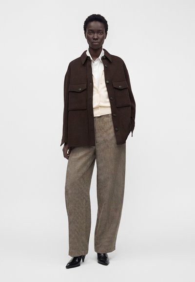 Femme portant une veste oversize marron foncé, un cardigan crème, une chemise blanche, un pantalon large à carreaux et des chaussures à talons noires, debout devant un fond uni.