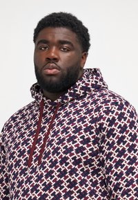 Hættetrøje med et geometrisk mønster i marineblå, mørkerød og cremegul, fremstillet af et tekstureret stof og med kontrasterende snore.