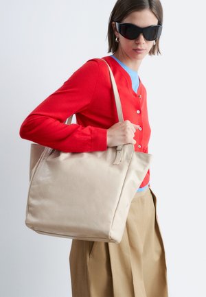 Vrouw in een rood vest en zonnebril, met een grote beige boodschappentas over de schouder, tegen een effen achtergrond.