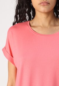Tricou roz cu mânecă scurtă și decolteu rotund, caracterizat printr-o textură moale și netedă și mâneci rulate pentru un detaliu suplimentar.