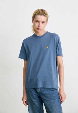 Giovane con i capelli biondi all'indietro che indossa una maglietta blu Carhartt e jeans blu su uno sfondo bianco semplice.