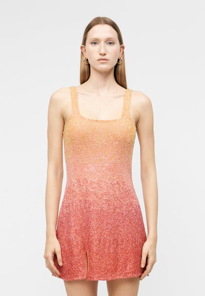 Jeune femme aux cheveux lisses portant une robe en maille dégradée orange à rose sans manches avec une petite fente à l'avant, debout devant un fond uni.