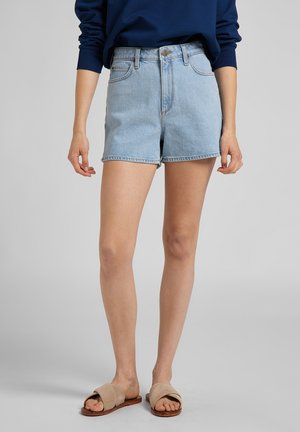 Mujer con shorts de mezclilla azul claro, camiseta de manga larga azul marino y sandalias beige sin talón, de pie frente a un fondo gris liso.
