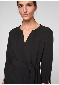 Robe noire avec un décolleté en V, des manches trois-quarts et une ceinture nouée à la taille. Le tissu semble lisse avec une texture subtile.
