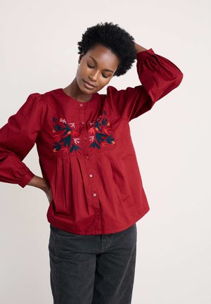 Rote Baumwollbluse mit Puffärmeln, Knopfverschluss und floralem Stickerei in Blau-, Rosa- und Orangetönen im Brustbereich.