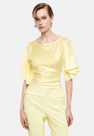 Donna che indossa una camicetta di raso giallo chiaro con maniche corte svasate e pantaloni gialli coordinati, in piedi contro uno sfondo grigio chiaro e uniforme.