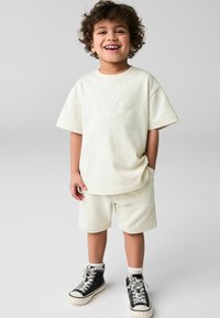 Lys beige bomulds t-shirt med korte ærmer, prydet med et cirkulært logo; sat sammen med matchende shorts og sorte høj-top sneakers.