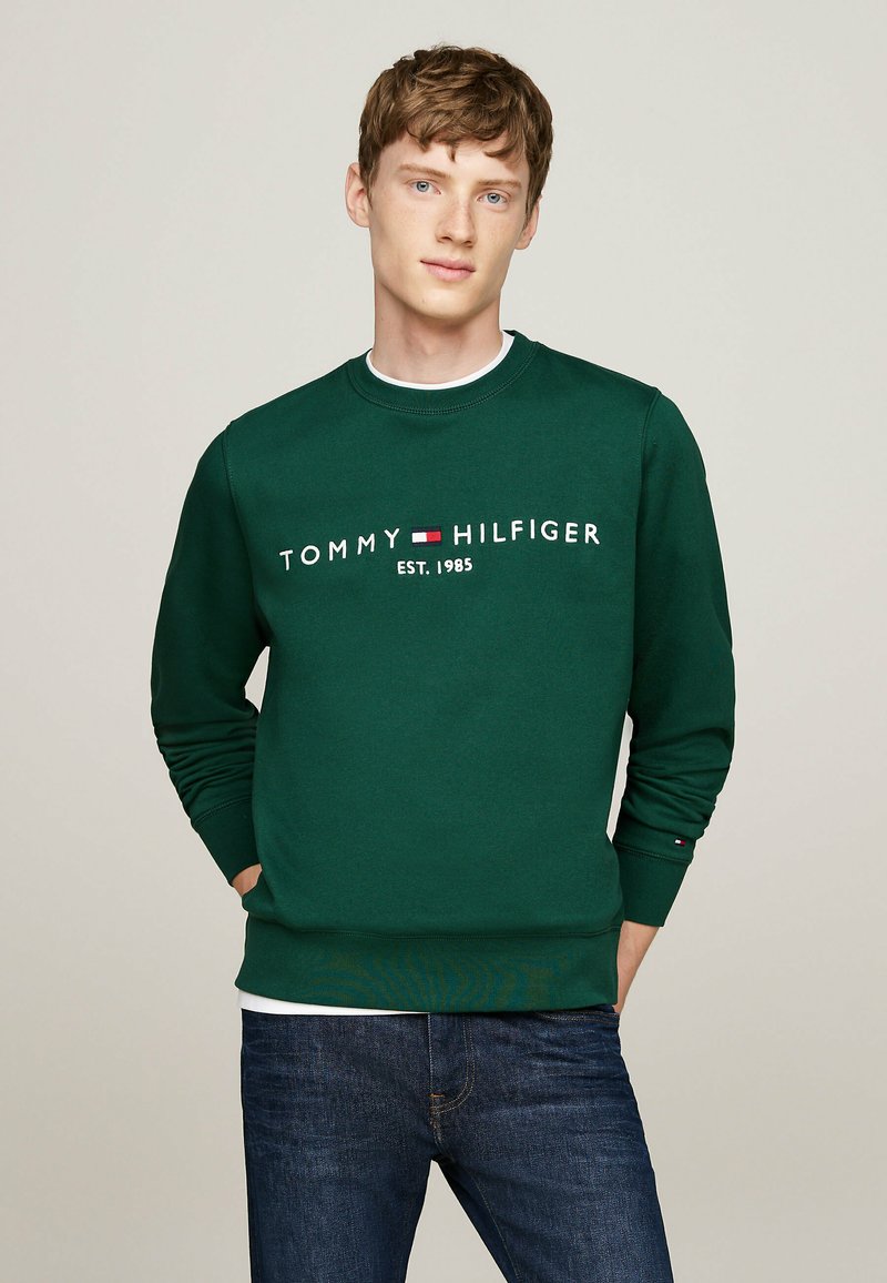 Un sweat-shirt vert à col rond en coton. Présente le logo blanc "TOMMY HILFIGER" et l'année "EST. 1985" au-dessus de la taille. Assorti avec un jean bleu.