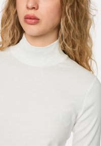 Weiße, figurbetonte Rollkragenbluse aus weichem Material mit geripptem Stoff und minimalen Nahtdetails, gezeigt von der Brust aufwärts.