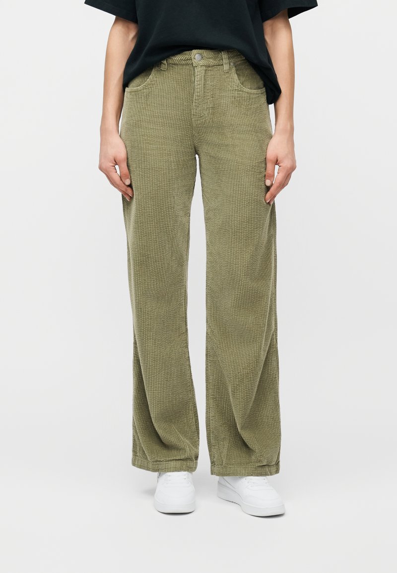 Άτομο που φορά φαρδιά βερμούδα από κοτλέ πράσινο ελιάς, μαύρο πουκάμισο και λευκά αθλητικά παπούτσια, στέκεται μπροστά σε απλό ανοιχτό φόντο.