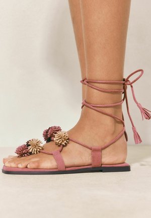 Picior purtând o sandale plată roz cu șireturi și decorațiuni cu pompoane pe barete, fotografiat pe un fundal neutru.