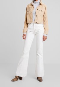 Veste en velours côtelé beige raccourcie, col roulé gris, jeans évasés blancs et bottines à talons carrés marron à motifs.